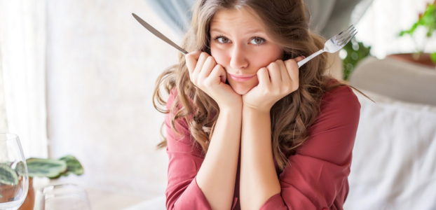 Woman with fork and knife in hands is sitting with her elbows on the table and a questioning look on her face.