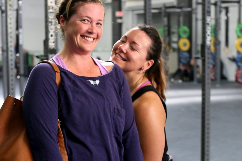 A female gym member smiles at the camera while her friend rests her smiling face on her shoulder.