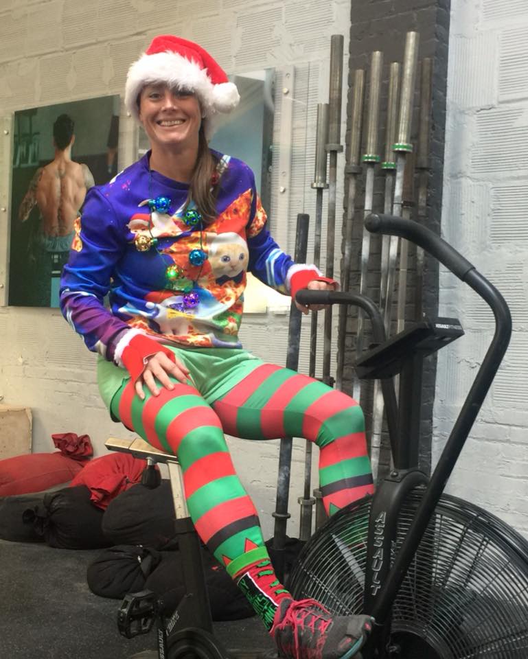 Invictus coach dressed in Santa hat, ugly sweater, and elf leggings sitting on the Assault Bike while waiting for her class to arrive.