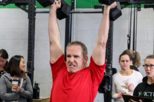 Male masters athlete holding two dumbbells overhead while others watch.