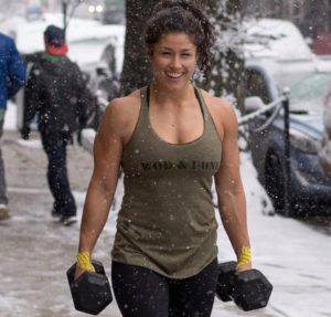 Female athlete doing farmer carries down a city sidewalk in the snow.