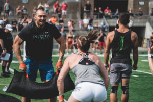 Male athlete doing a sandbag clean in competition while his female teammate cheers him on.