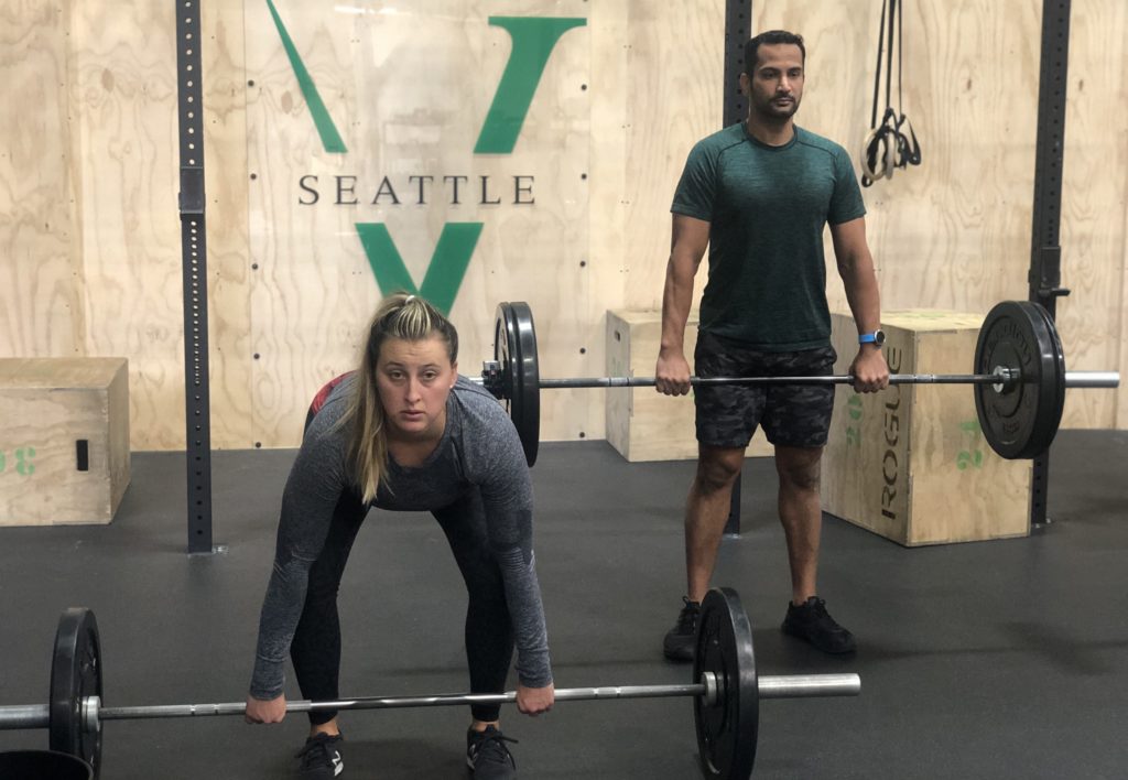 A male athlete at the top of a deadlift in the background with a female athlete pulling a deadlift from the floor.
