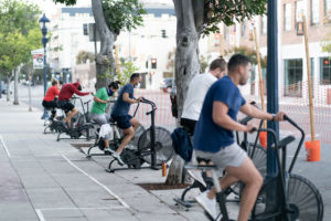 Group of athletes riding Assault Bikes on the sidewalk outside of the gym.