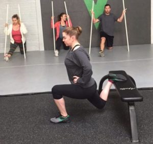 Female athlete doing Bulgarian Split Squat with hands on hips while three athletes watch while stretching.
