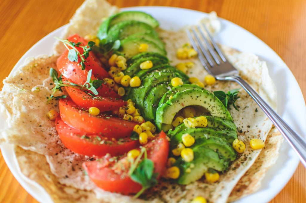 Open face tortilla on a plate topped with sliced tomatoes, avocado, corn and sprouts.