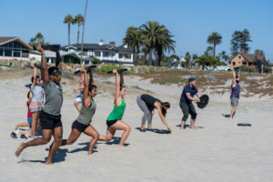 Group of athletes at the beach doing overhead plate hold lunges in a line.