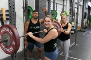 Three athletes standing around their racked barbell and smiling for the camera.