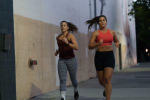 Two female athletes running on a sidewalk around the gym.