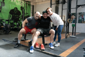 Three athletes huddled together on a weight bench smiling for the camera.