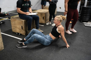 Female athlete sits on the ground after a workout while her competition judge sits on a box and tallies her reps.