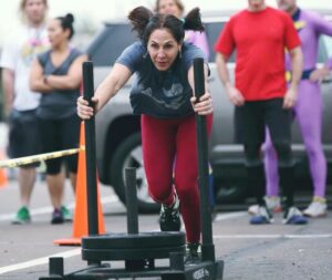 Female Masters athlete does sled pushes outside the gym while other athletes anxiously await their turns.
